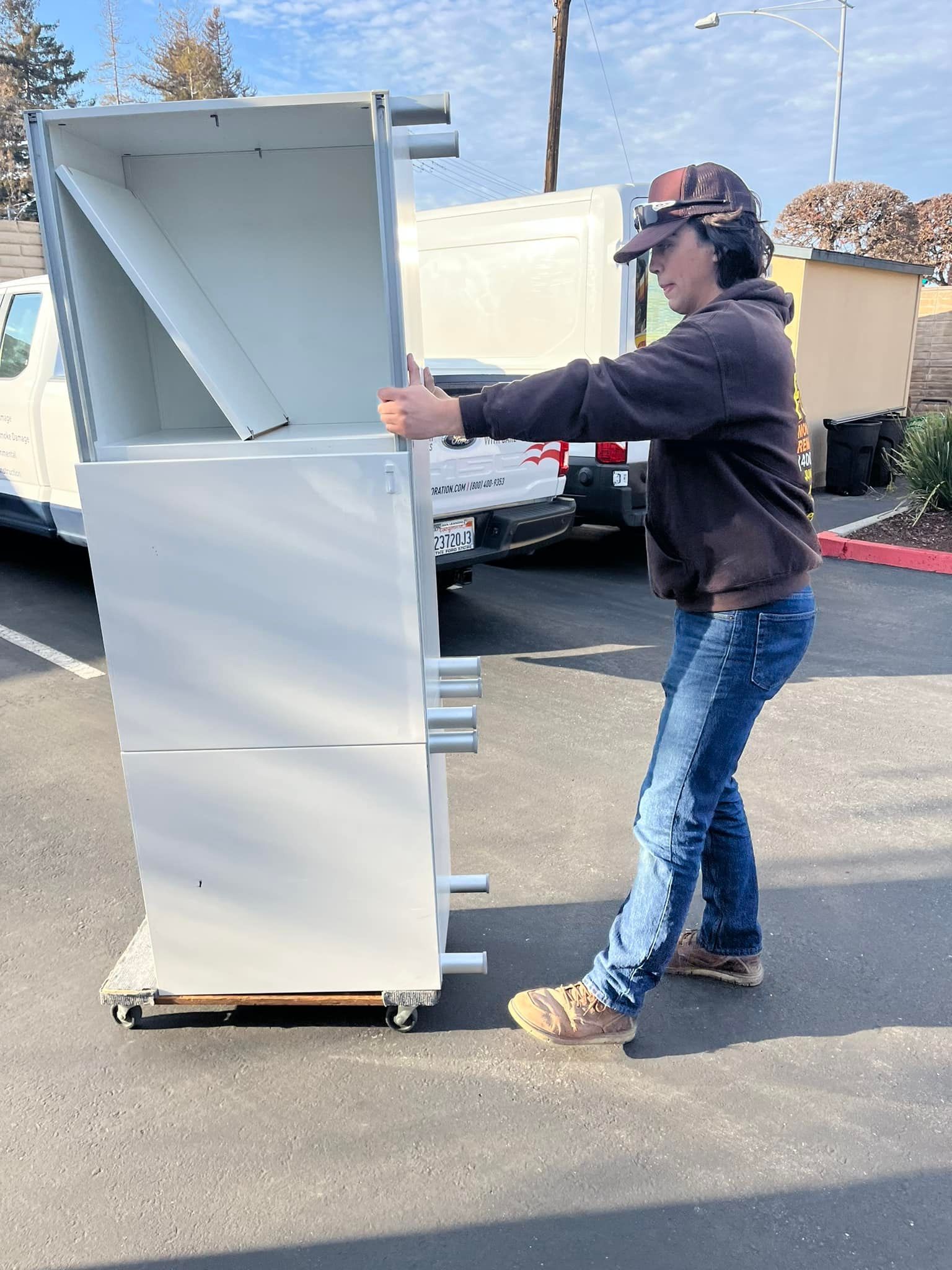 Person in jeans and brown shirt maneuvering a large, white cabinet on a wheeled platform outside.