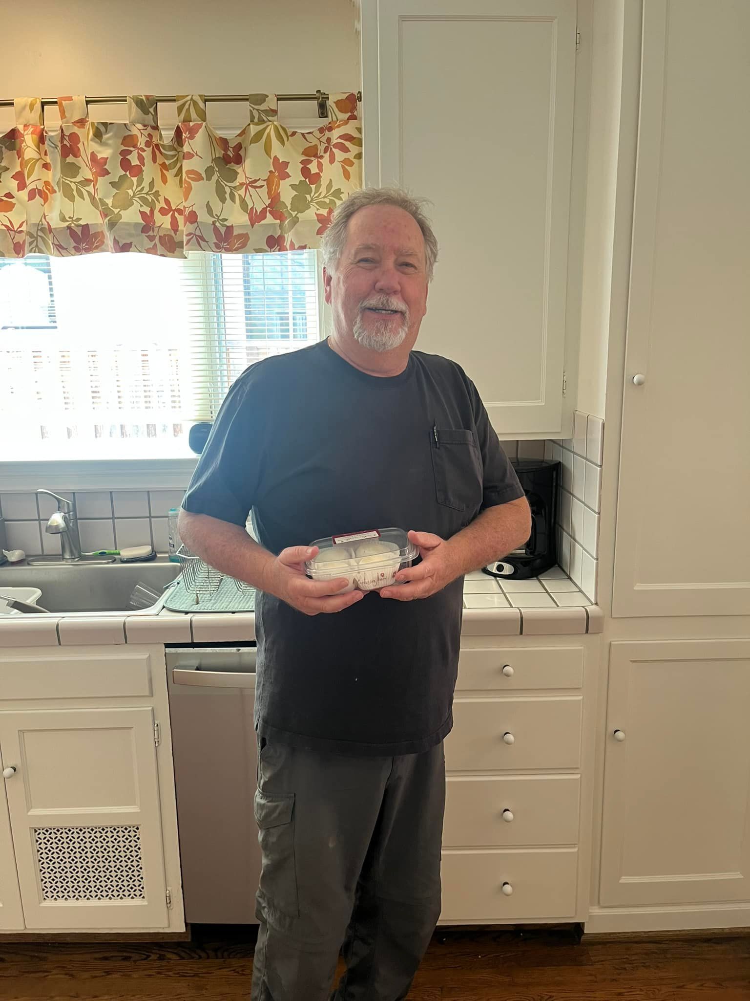 Man holding a rectangular object in a kitchen with white cabinets, near a window with floral curtains.