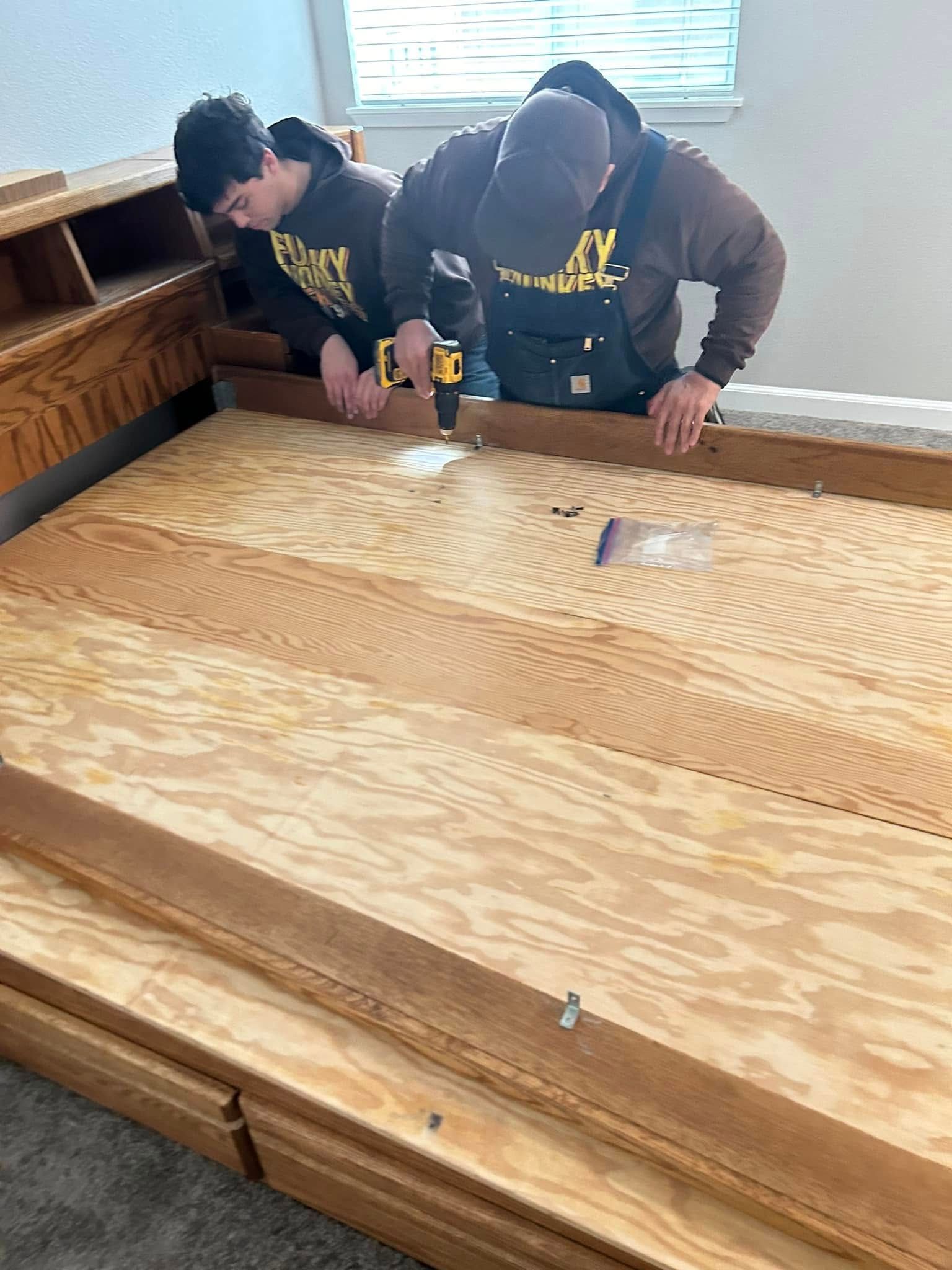 Two people assembling a wooden bed frame, one using a drill.
