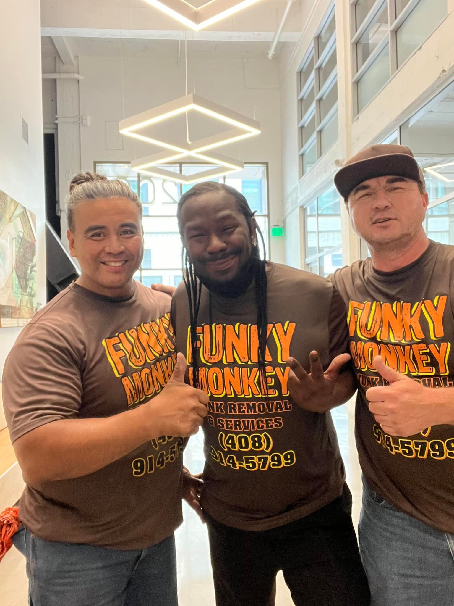 Three people in brown shirts posing, inside modern space. Shirts say 