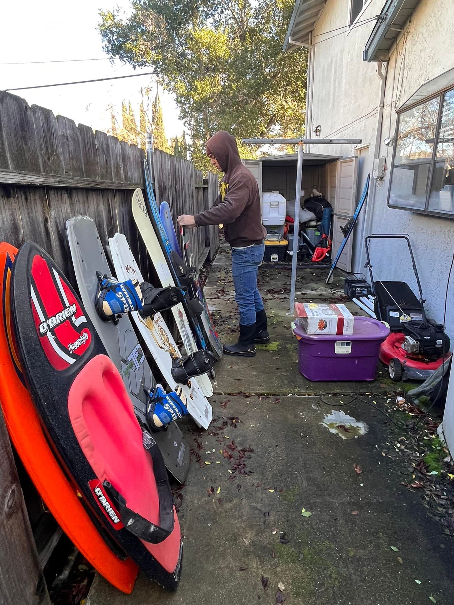 Person in hooded sweatshirt examining snow sleds leaning against a wooden fence.