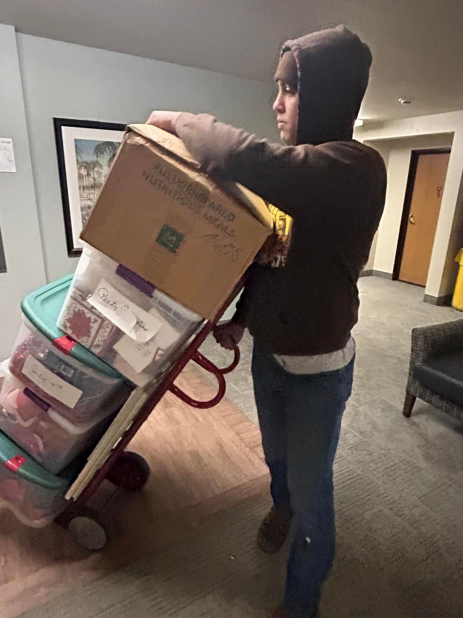Person in a brown hoodie pulls a hand truck loaded with boxes and bins in a hallway.