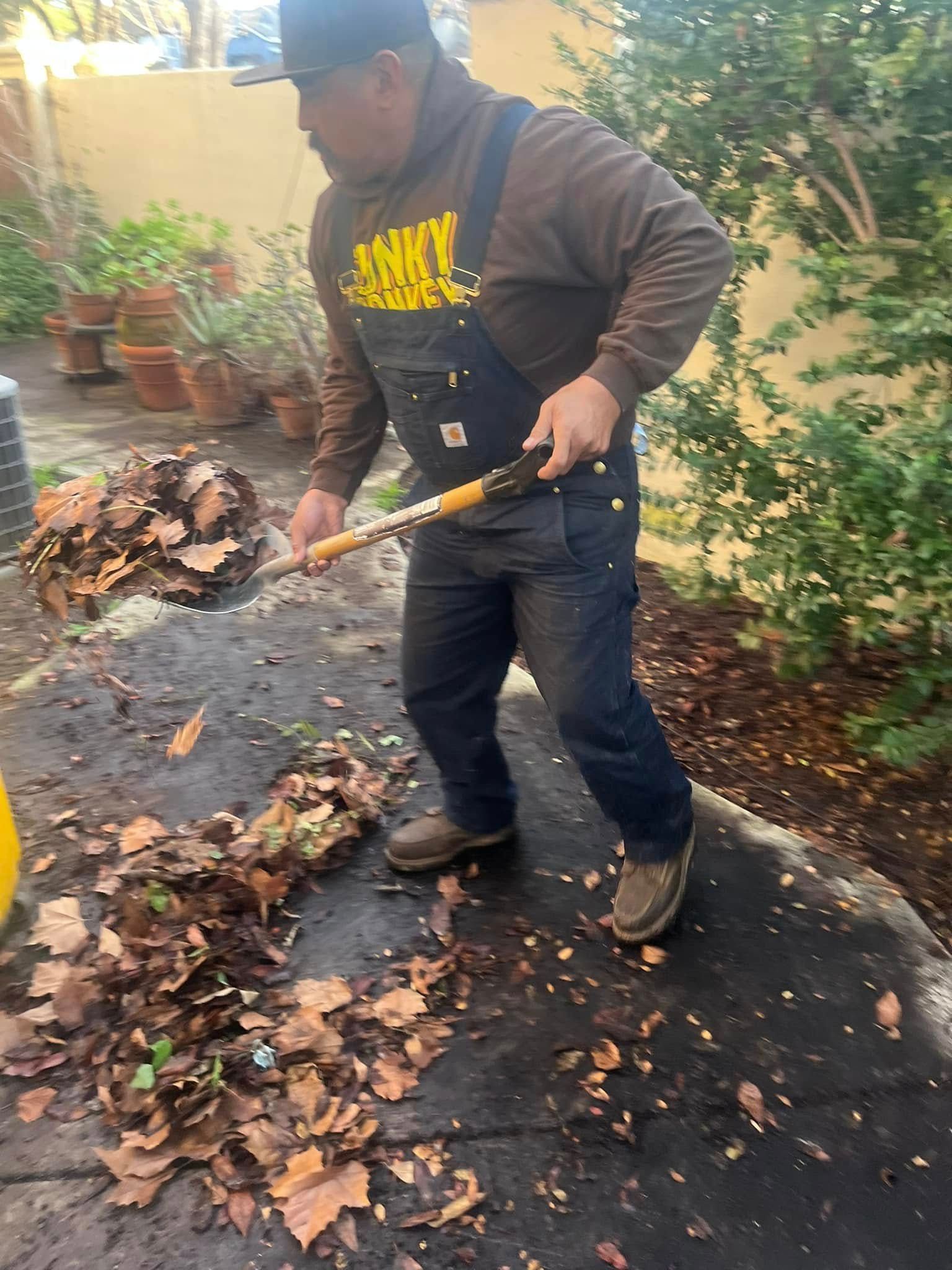 Man in overalls shoveling leaves on a paved patio; potted plants in the background.