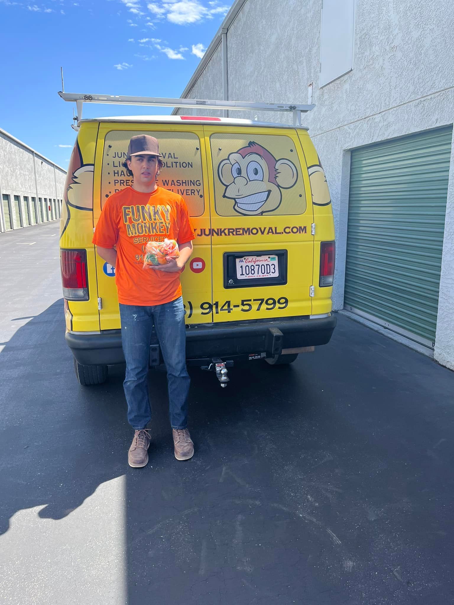 Man in orange shirt holds package in front of yellow van with logo, parked outside.