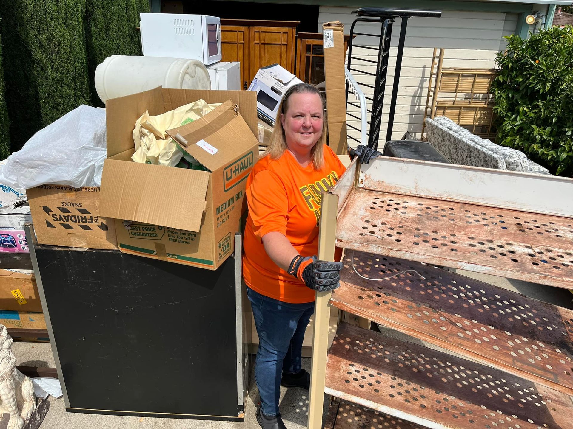 Woman in orange shirt, gloved, stands next to pile of discarded furniture, holding a metal frame.