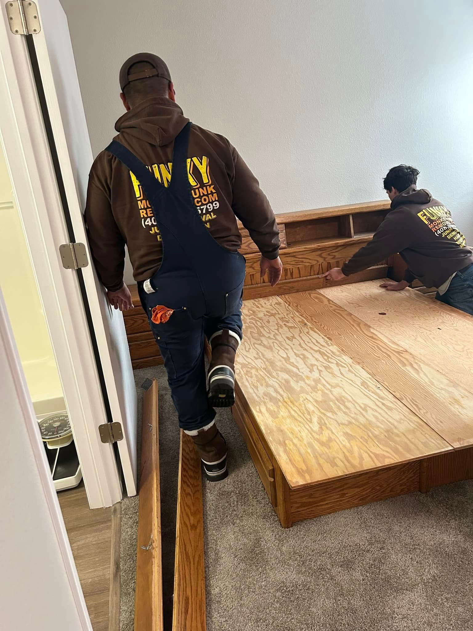 Two people move a wooden bed frame through a doorway. One person walks, other adjusts the frame.