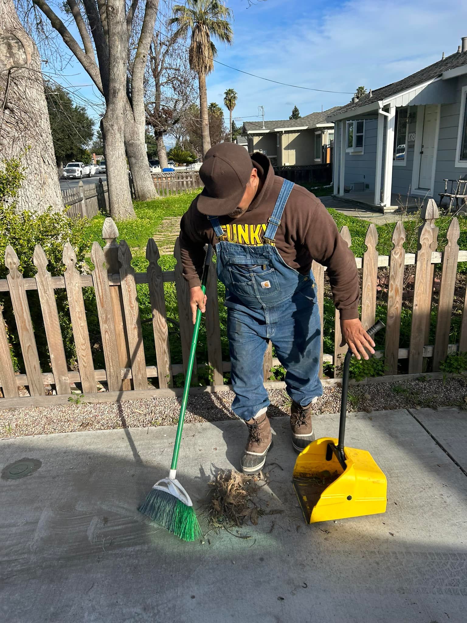 Person sweeping debris into a dustpan outside by a fence and houses on a sunny day.