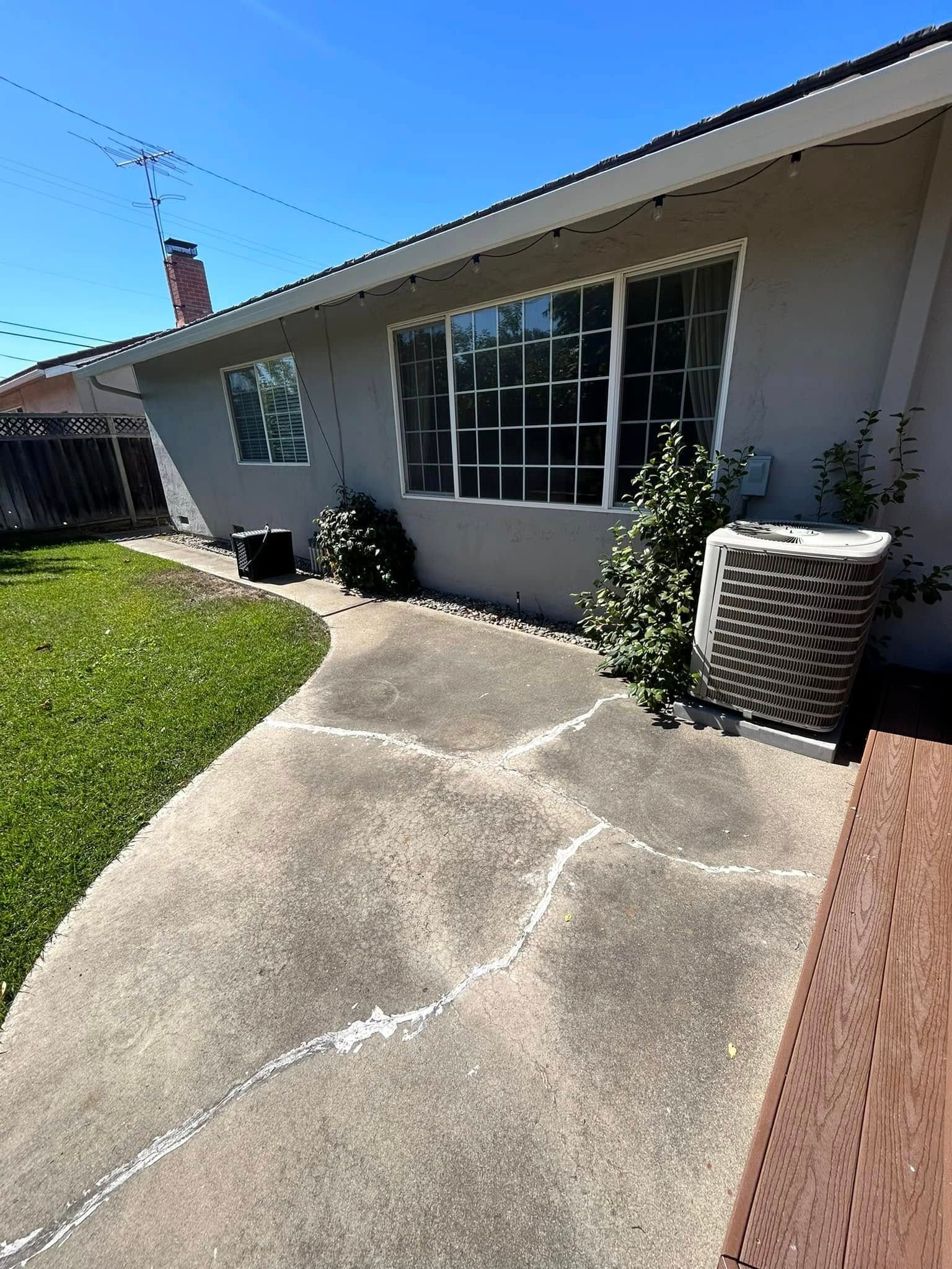 Concrete path alongside a light-colored house with a large window and air conditioning units; green grass and blue sky.