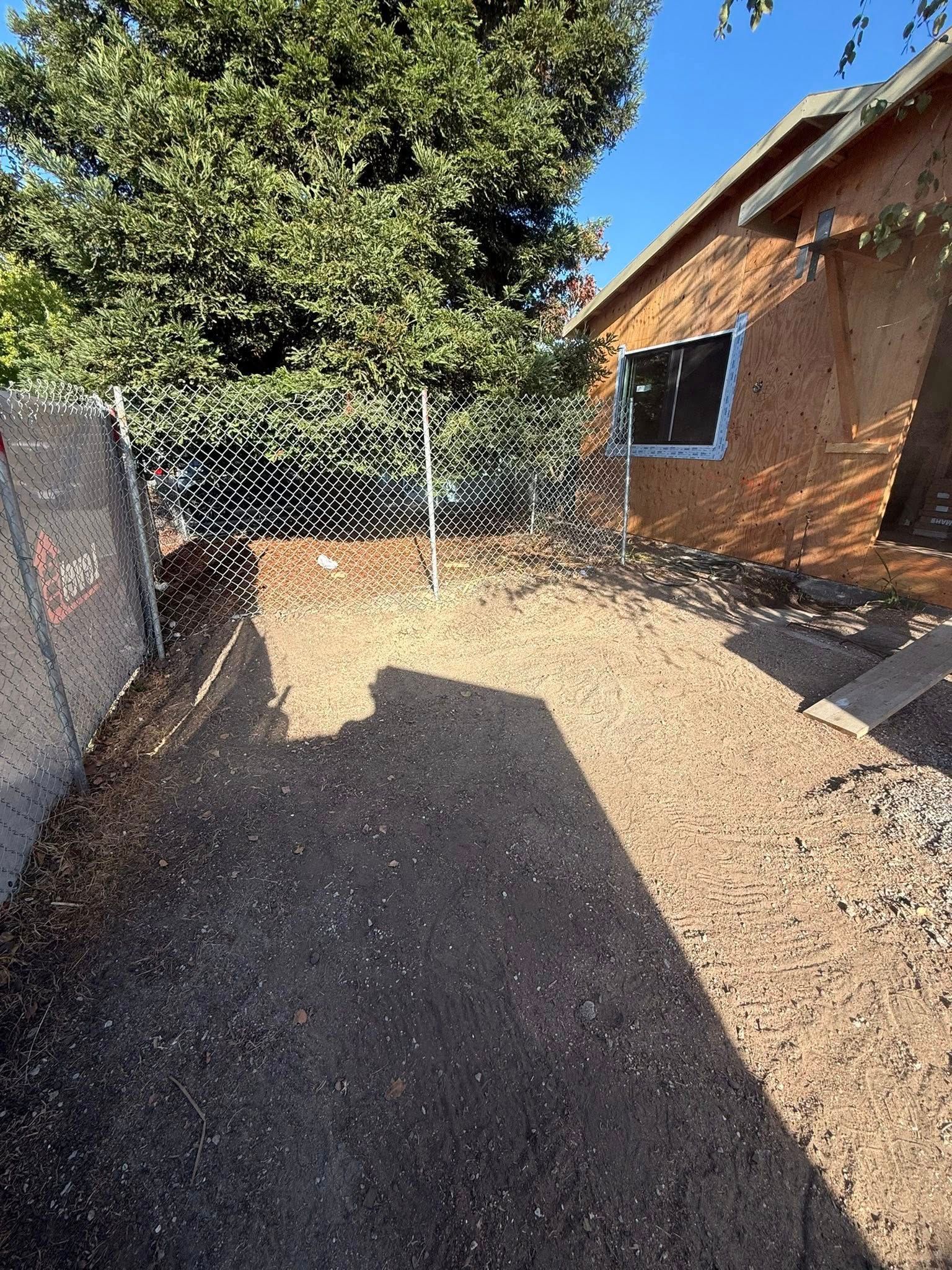 Dirt yard enclosed by a chain link fence with a building in the background and shadow from a tree.