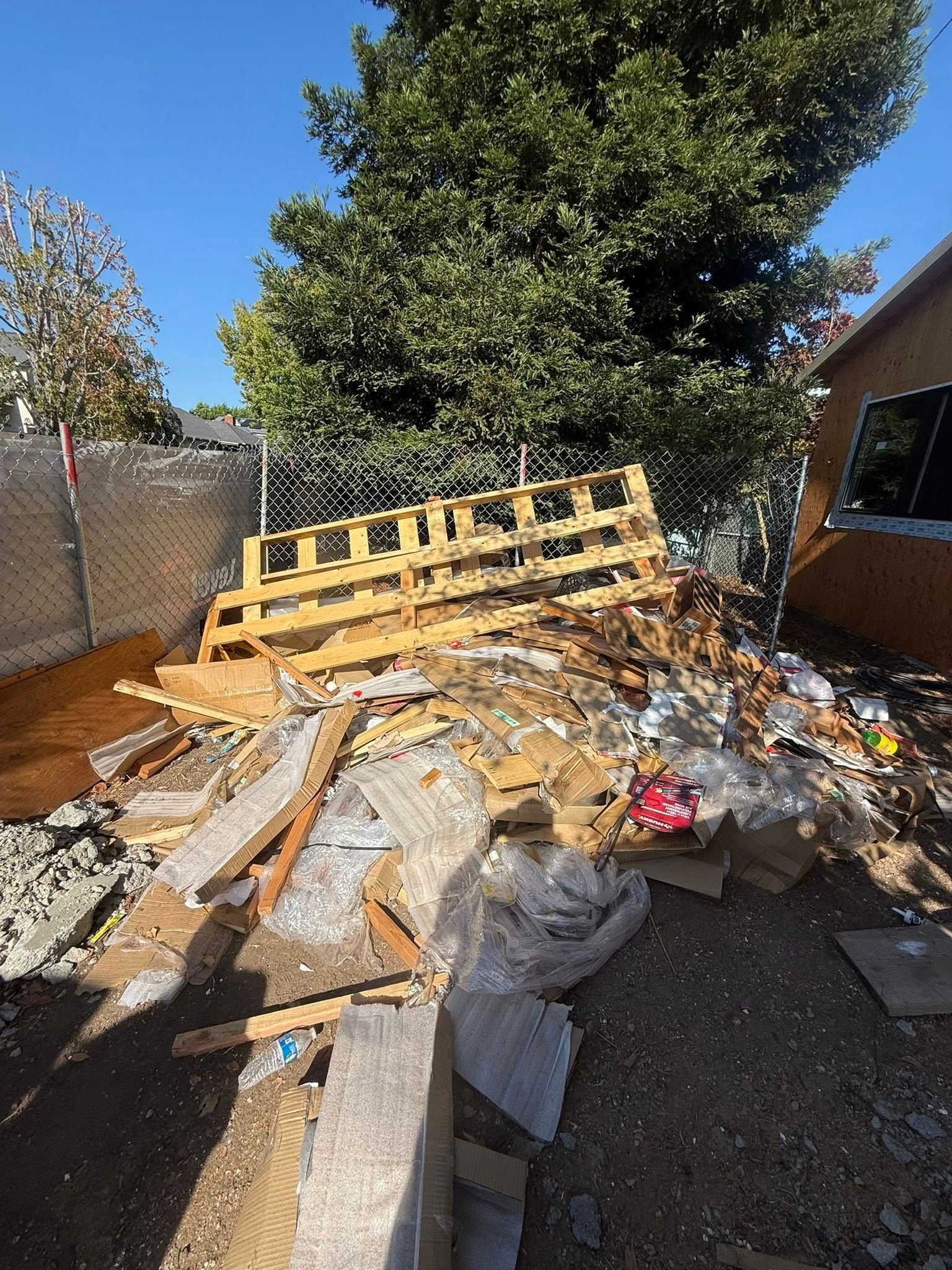 Pile of construction debris and wood pallets near a fence and a tree against a clear sky.