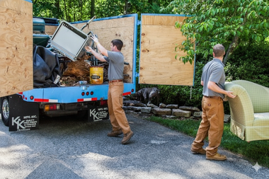 Two movers unloading a truck, one with a chair cushion, the other with a metal appliance; outdoors.