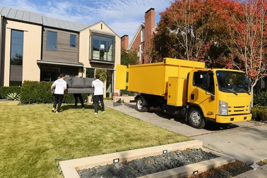 Movers carrying a couch to a yellow truck parked in front of a modern house.