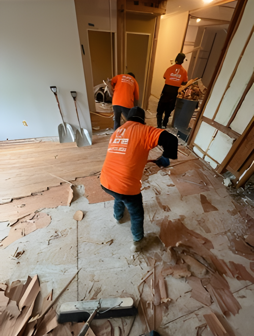 Three workers in orange shirts demolishing a room. Shovels lean against a wall. Debris covers the floor.
