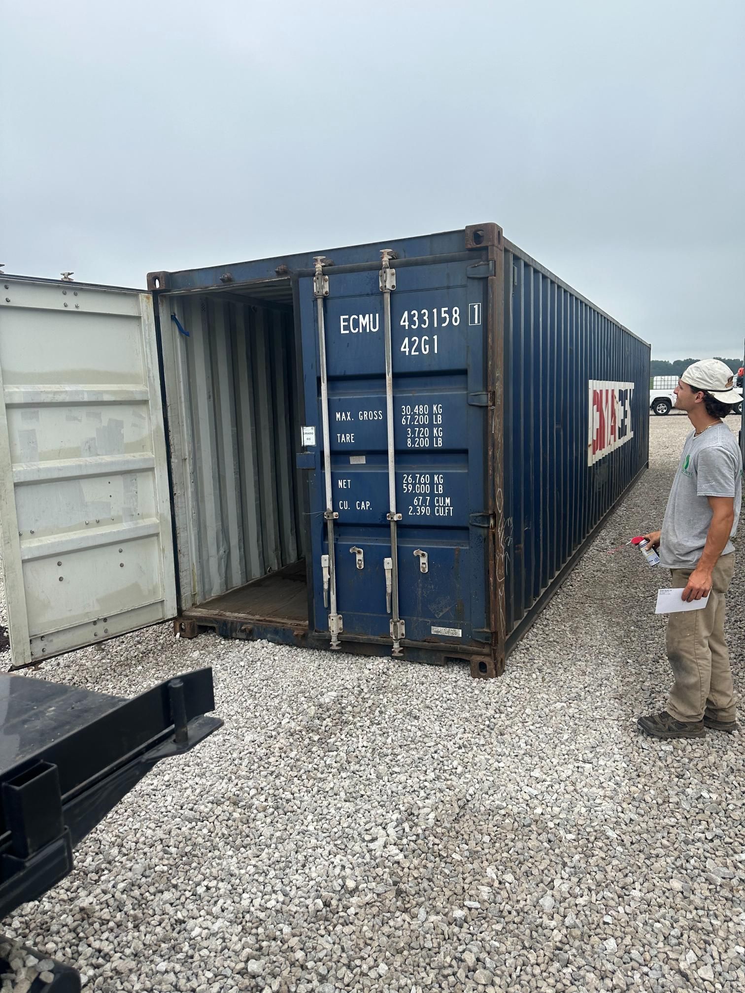A man is standing next to a blue shipping container in a gravel lot.