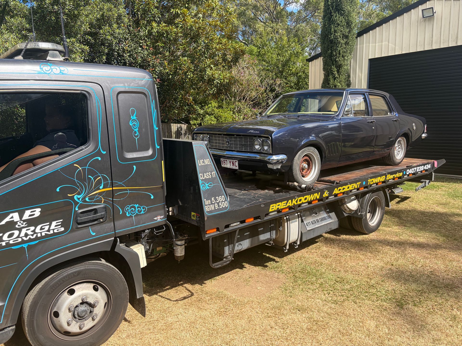 Tow truck carrying a black classic car on a flatbed beside a green shed — A B & Forge Towing In Caboolture, QLD