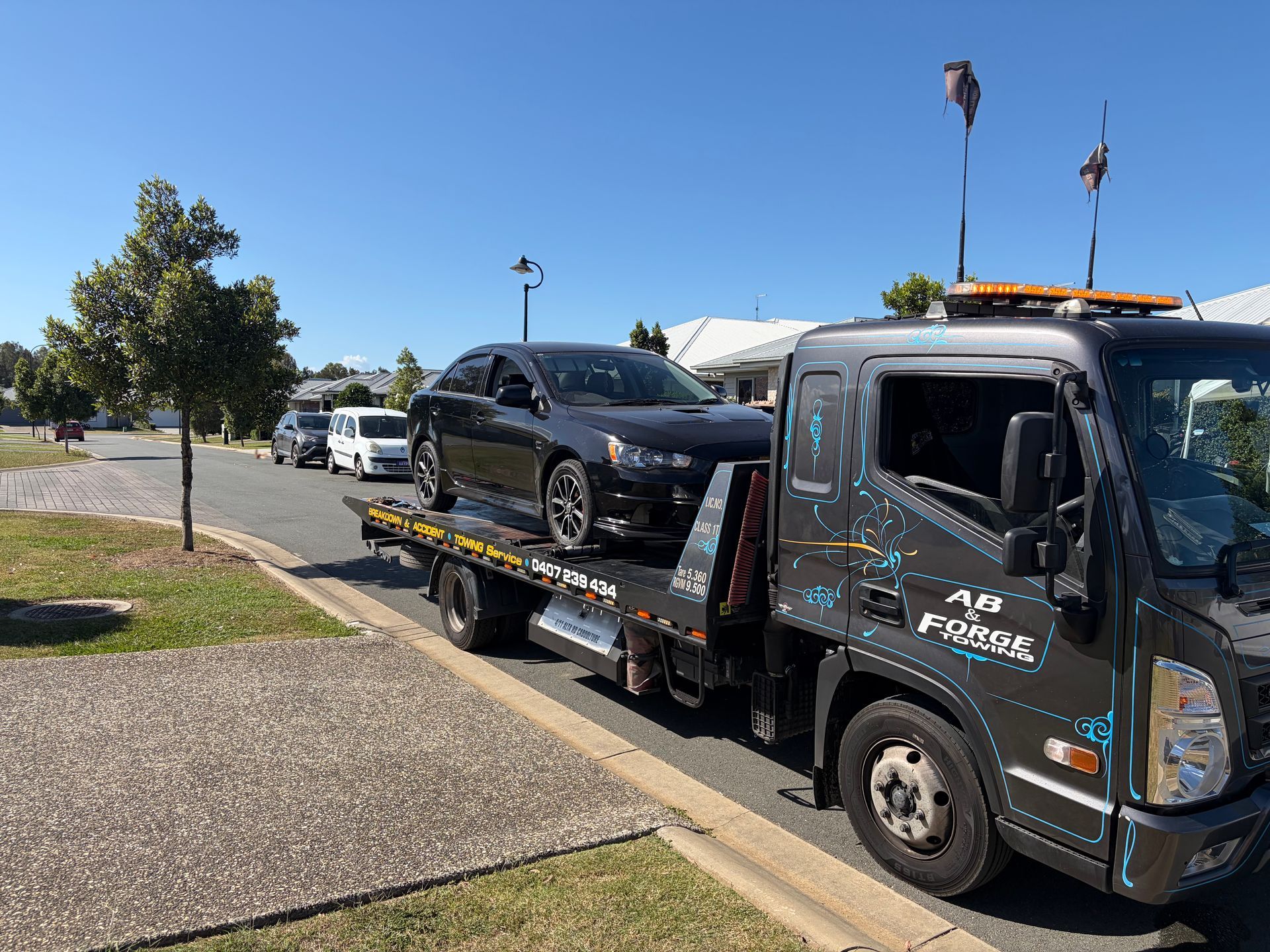 Tow truck hauling a black sedan on a flatbed along a sunny suburban street — A B & Forge Towing In Caboolture, QLD
