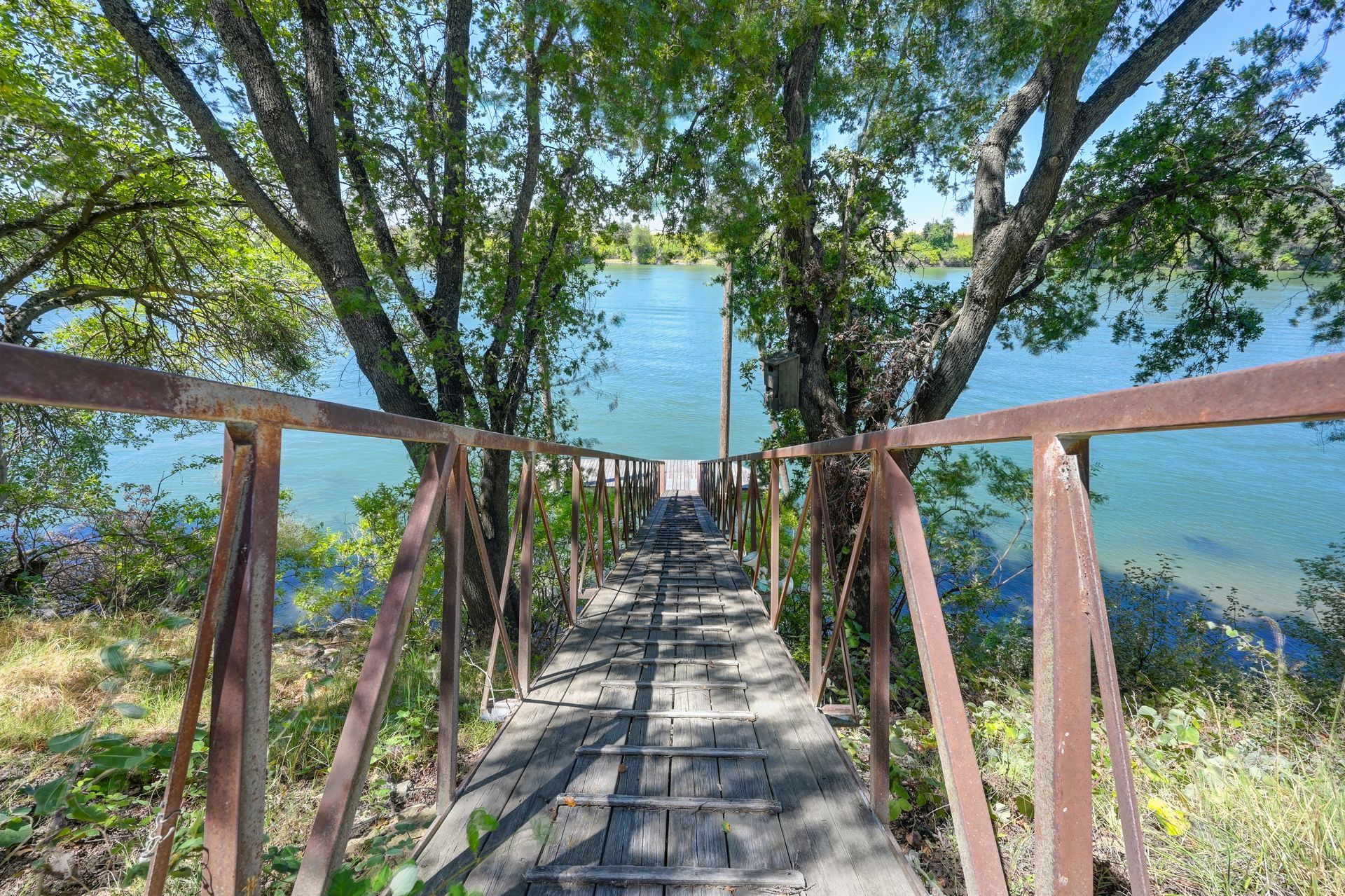 Wooden dock with rusty metal railings leading down to a calm, blue river surrounded by lush green trees on a sunny day.