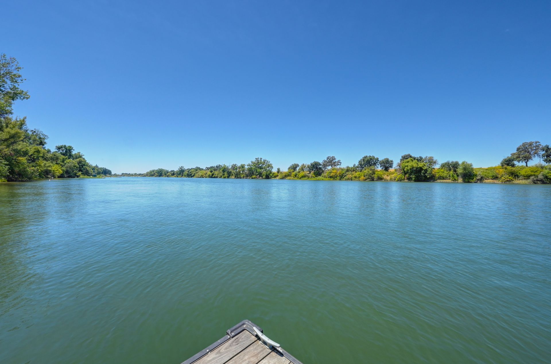 View of a river under a clear blue sky, seen from the edge of a wooden dock with greenery along the distant shoreline.