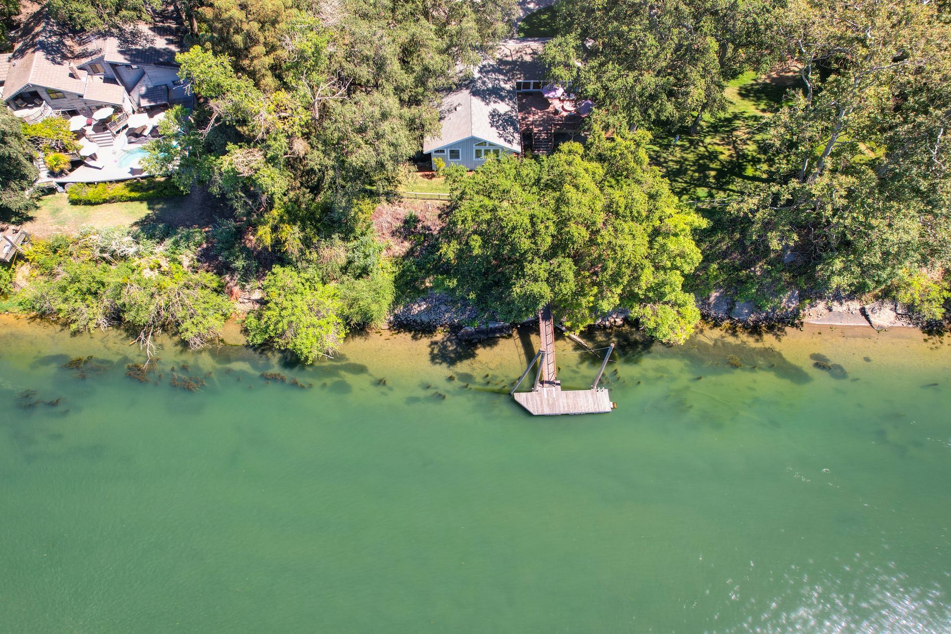 Aerial view of a riverside home surrounded by trees with a private wooden dock extending over clear, green water.