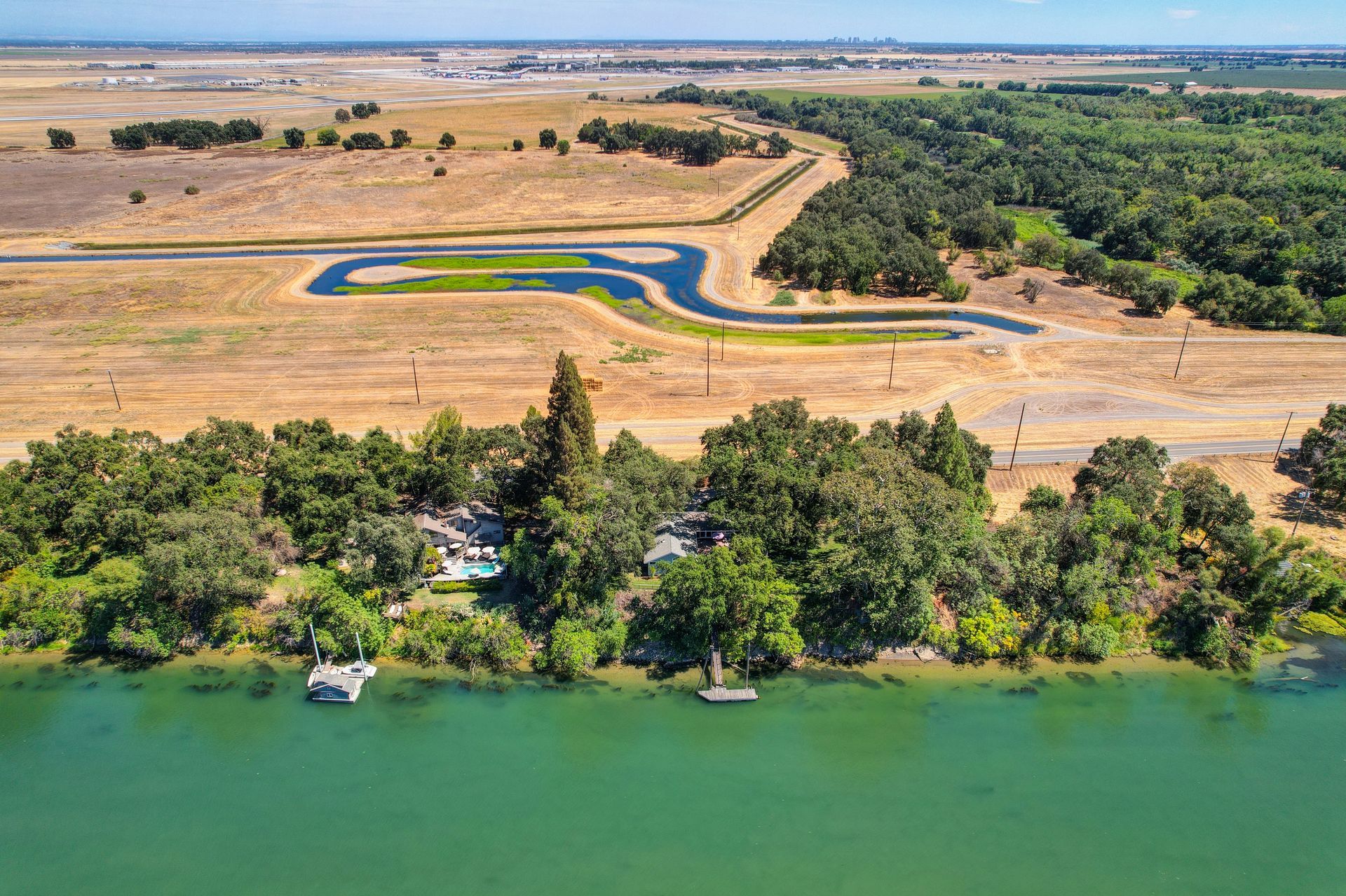 Aerial view of a riverside home with a private dock and pool, surrounded by trees and open fields under a blue sky.