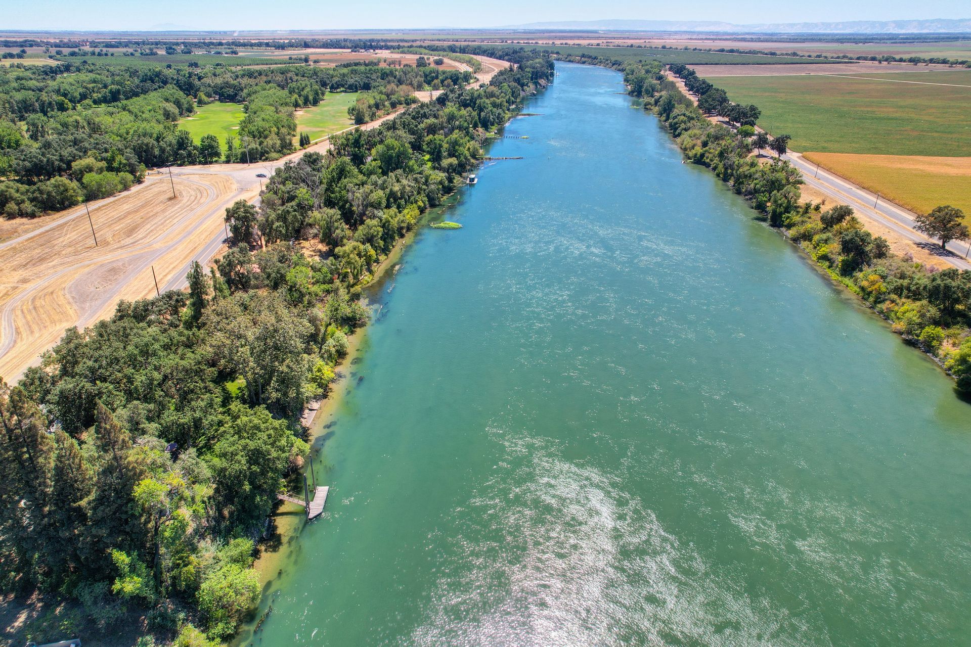 Aerial view of a wide river bordered by lush trees and open fields under a blue sky.