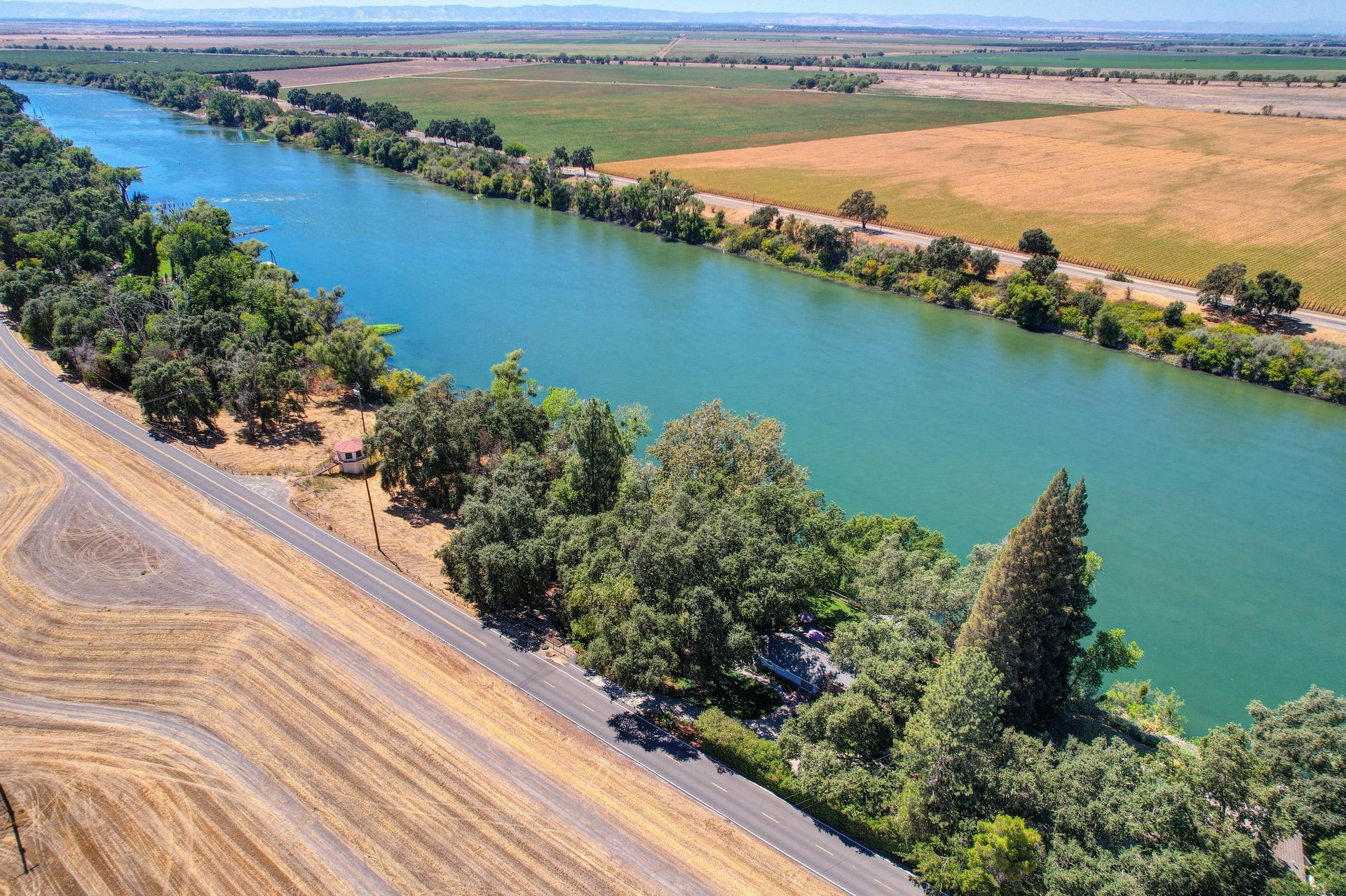Aerial view of a river bordered by lush trees and expansive farmland, with a narrow road running alongside the water.