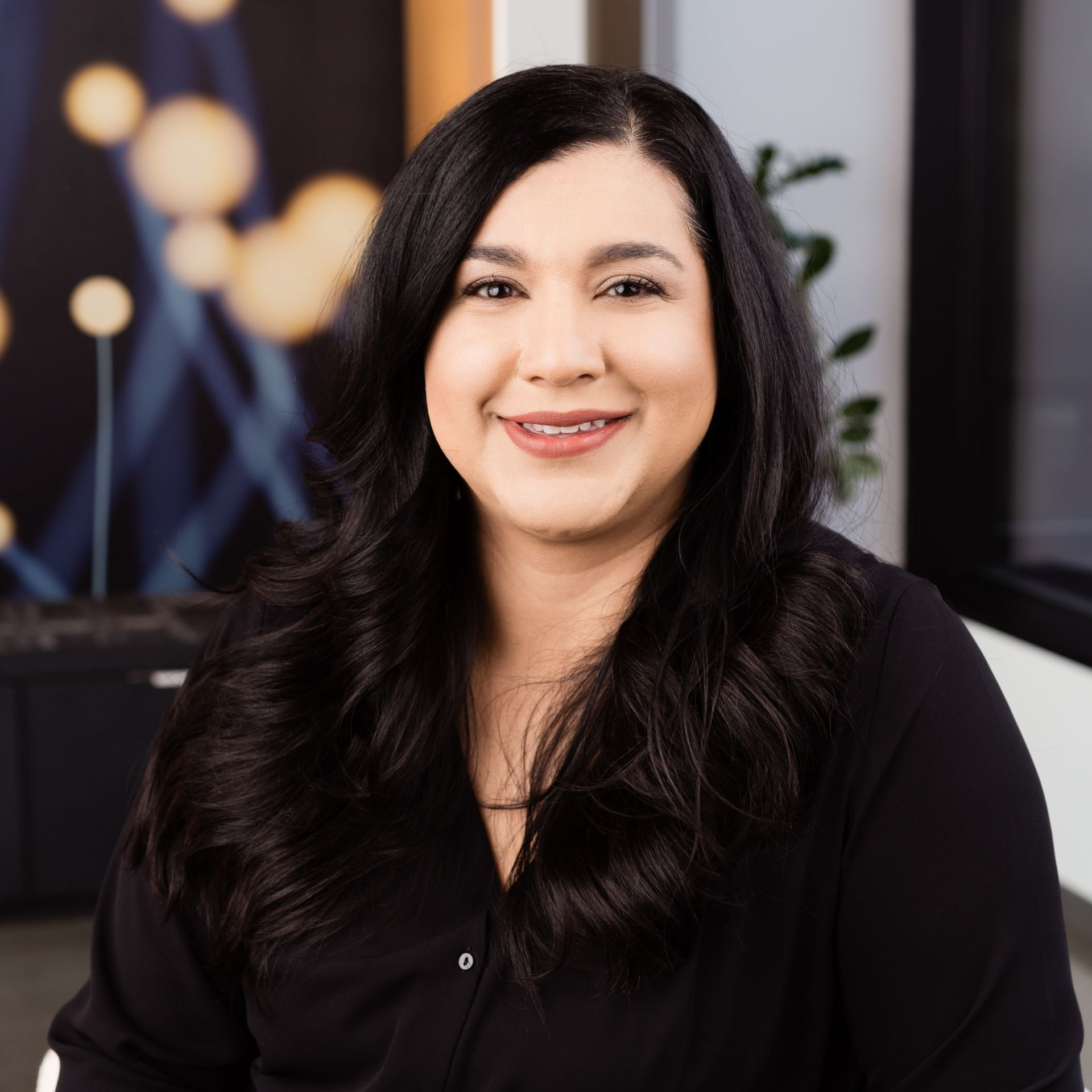 Woman with long dark hair, smiling, wearing a black shirt, in an office setting.