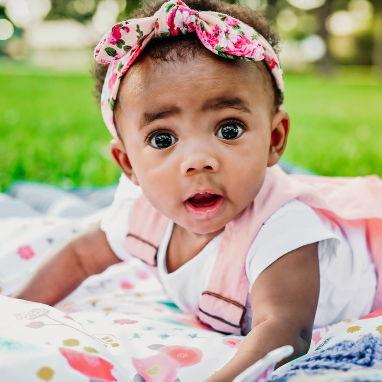 Baby lying on a blanket, wearing a pink floral headband and dress, mouth open, looking forward.