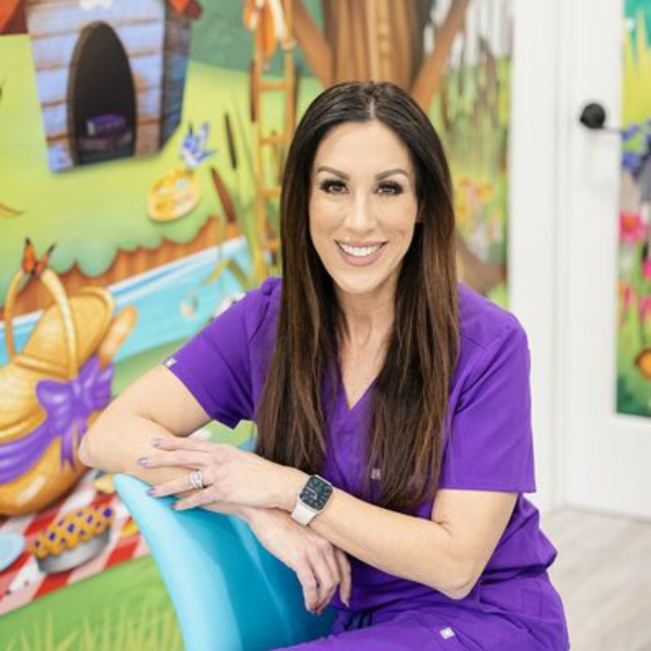 Woman in purple scrubs smiles, sits in a blue chair, colorful cartoon mural backdrop.