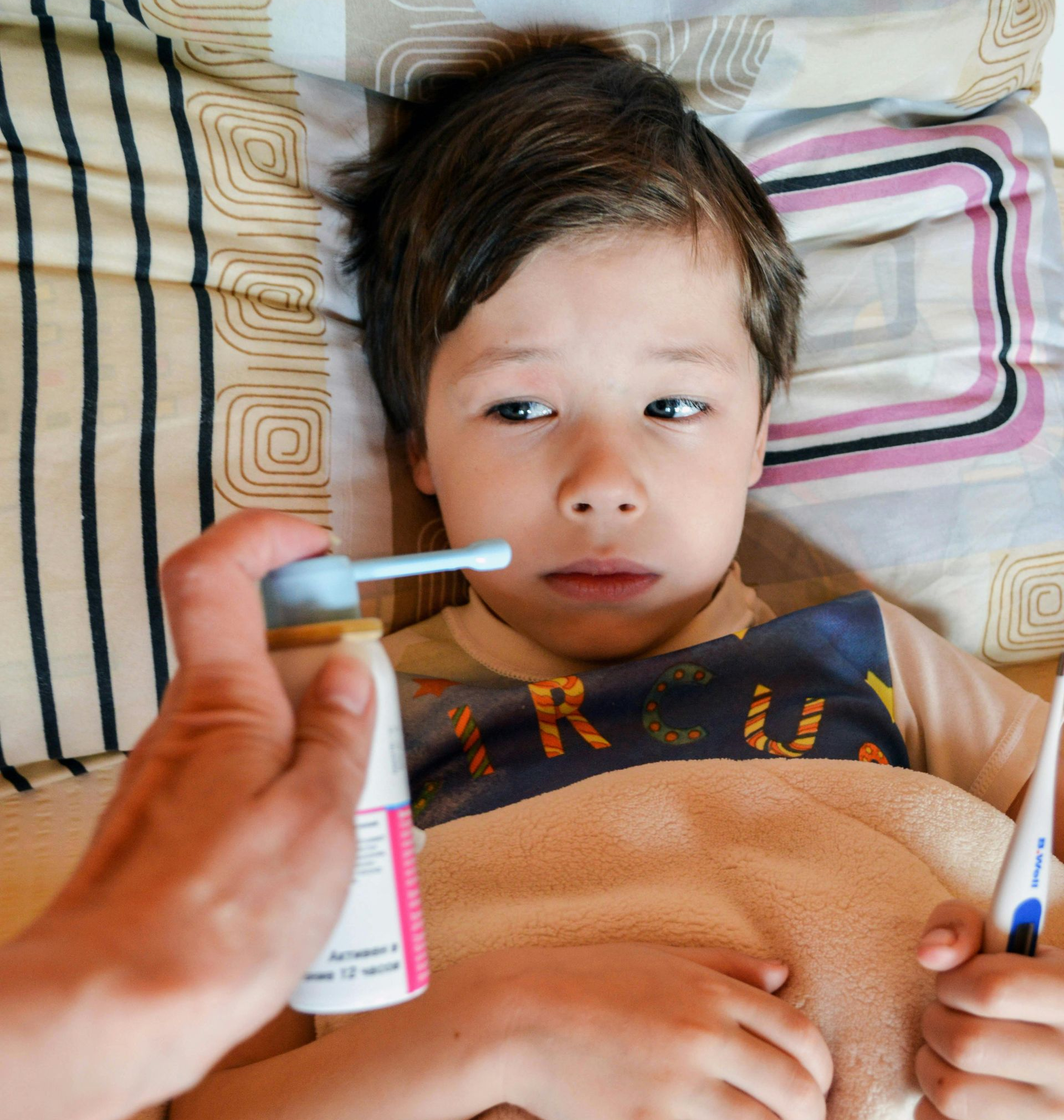 Woman assisting child with blowing nose. Both are in pajamas, indoors on a bed.
