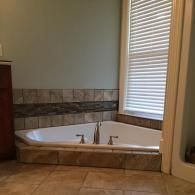 A white corner soaking tub with a central faucet, tiled surround, and beige floor tiles in a bathroom next to a window.