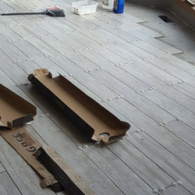 Light-colored, wood-look floor tiles being installed with spacers, with cardboard packaging pieces lying on top.