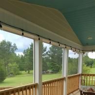 A screened-in porch with a teal ceiling, white railing, and string lights overlooking a grassy lawn and pine trees.