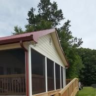 A tan screened-in porch with a red metal roof and a wooden wheelchair ramp, set against a backdrop of green trees.