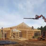 Construction workers frame a house structure under a blue sky, using a red telehandler to hoist lumber.