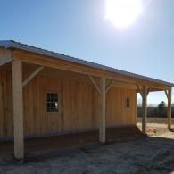 A rustic wooden barn featuring a covered porch with supporting beams, set against a bright, sunny sky.