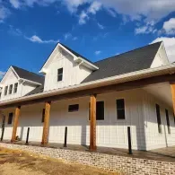 A white farmhouse with a dark roof and a wide, covered wooden porch, set against a bright blue, partly cloudy sky.