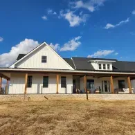A modern white farmhouse with a large covered porch, dark roof, and wood accents under a bright blue sky.