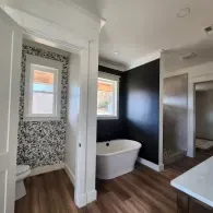 A bathroom with wood-look floors, a white soaking tub, and an accent wall with patterned wallpaper and a black finish.