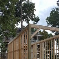 A wooden pergola or trellis structure under construction, with a multi-story house and trees in the background.