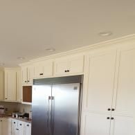 A stainless steel refrigerator sits between cream-colored kitchen cabinets with crown molding under recessed ceiling lights.