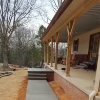 A covered wooden porch with structural beams, a concrete walkway, and brick foundation steps leading to a yard with trees.
