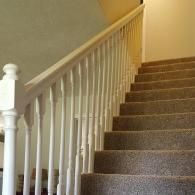 A staircase with brown carpeted steps leading upward, bordered by a white wooden railing and balusters.