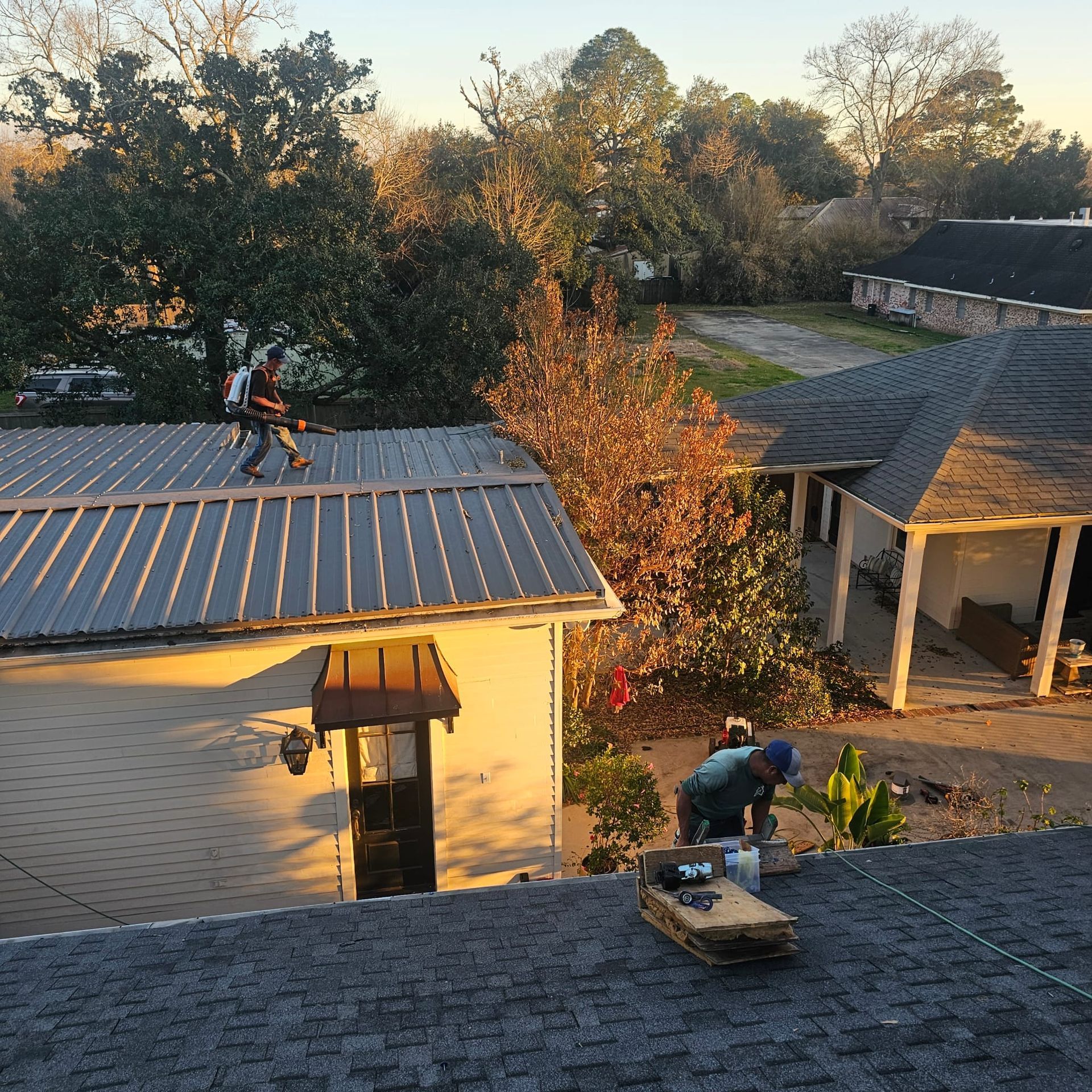 Workers on rooftops, installing metal roofing. Sunlight, trees, and other buildings are visible.