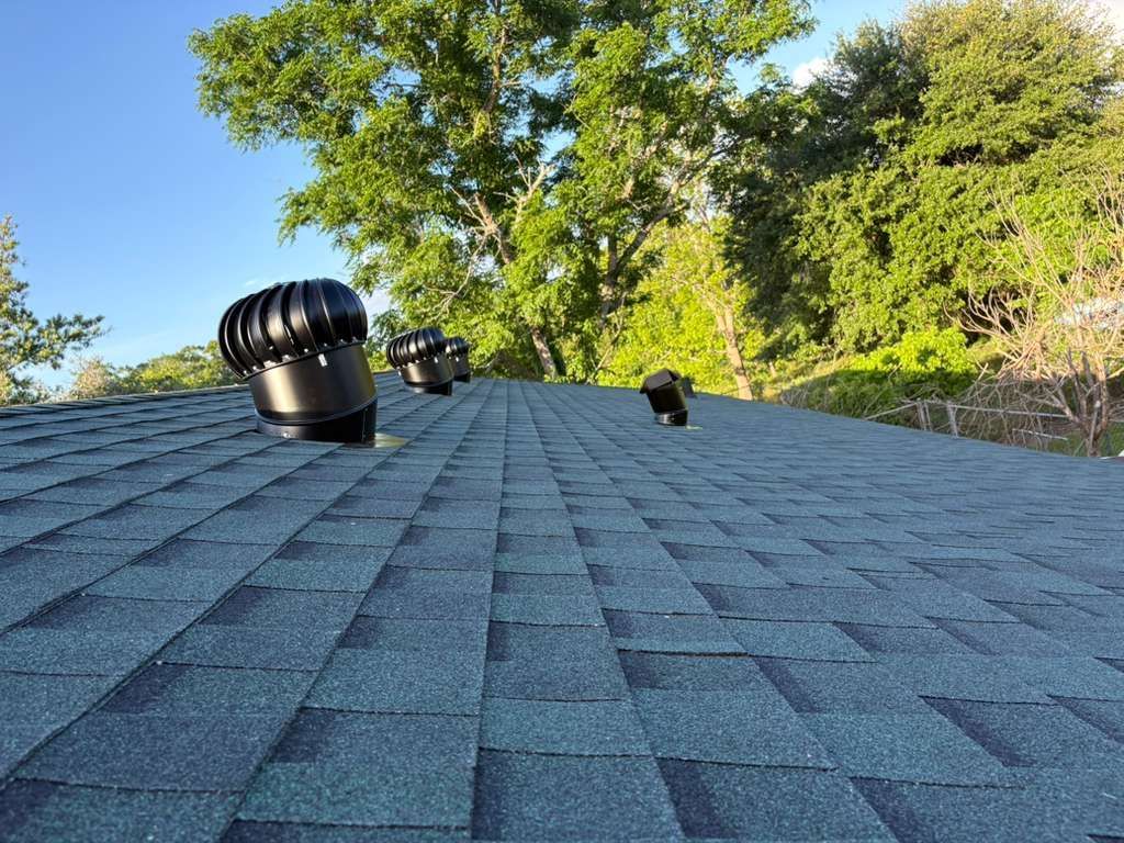 Blue shingled roof with two black vents and leafy trees against a clear sky