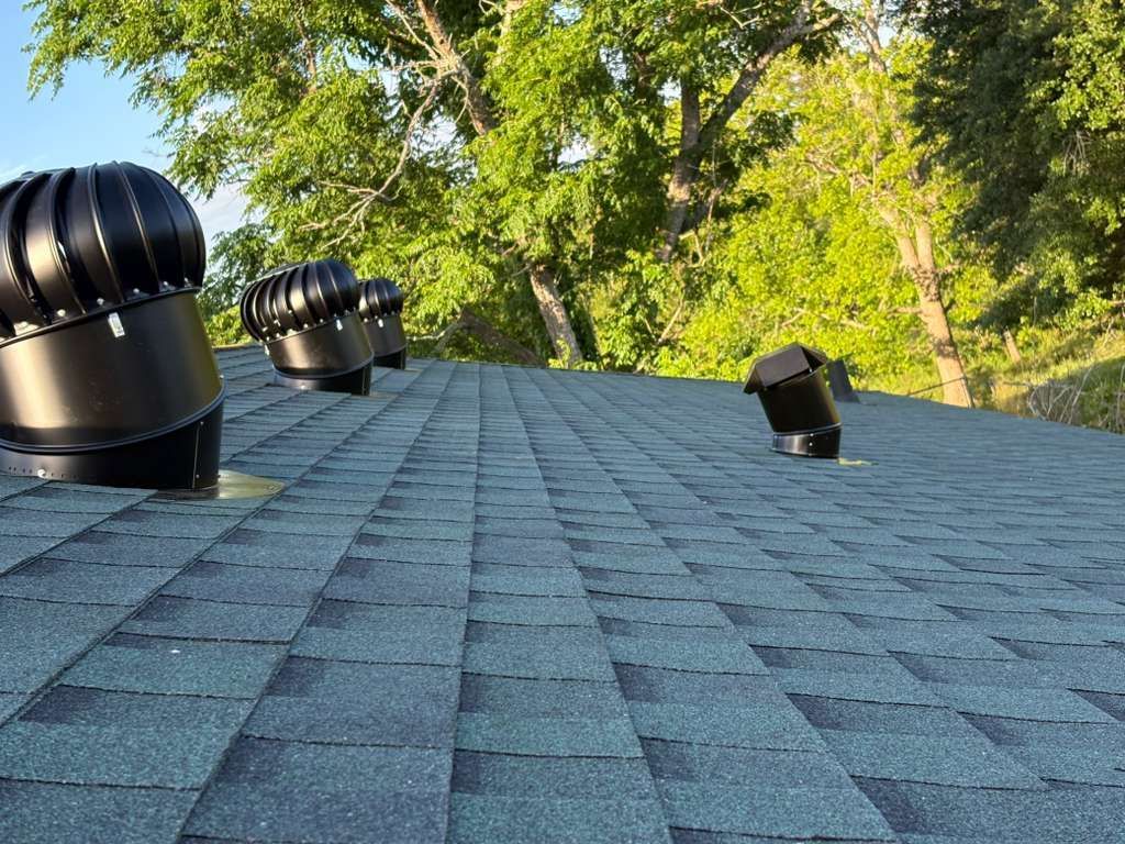 Blue shingled roof with two black vents and trees in the background
