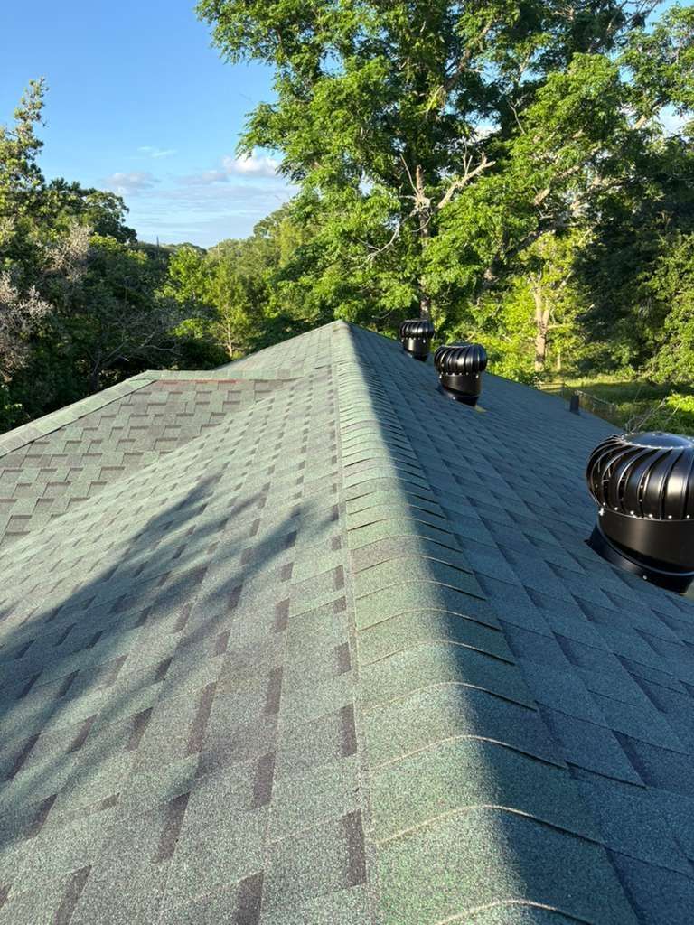 Roof shingles on a house with two black vents, viewed along the ridge line amid green trees and blue sky