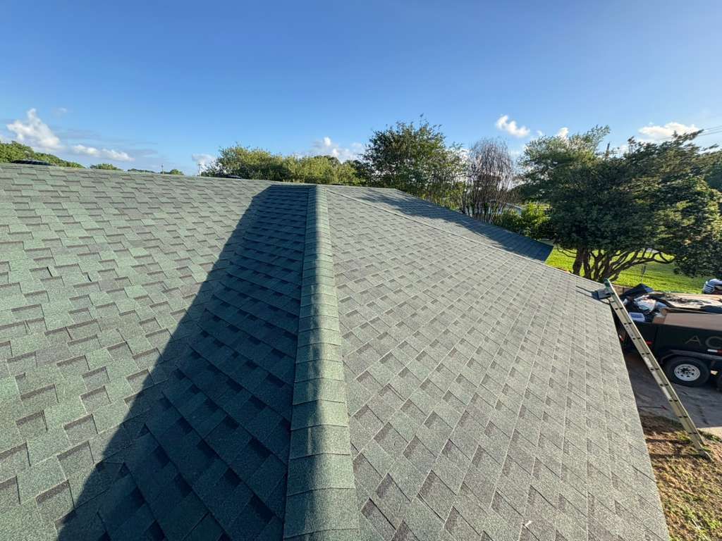 Green shingled roof with a central ridge under a blue sky, trees and parked vehicles in the background