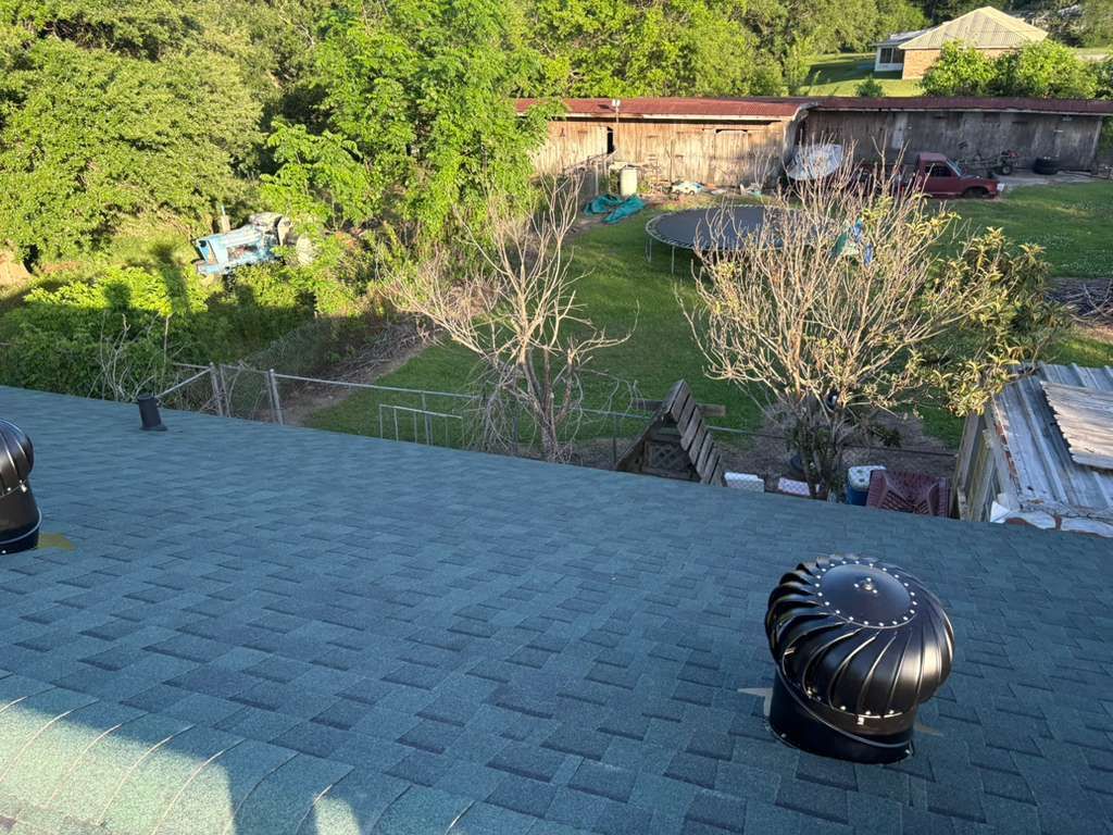 Rooftop shingles with black vents overlooking a sunny backyard and trees