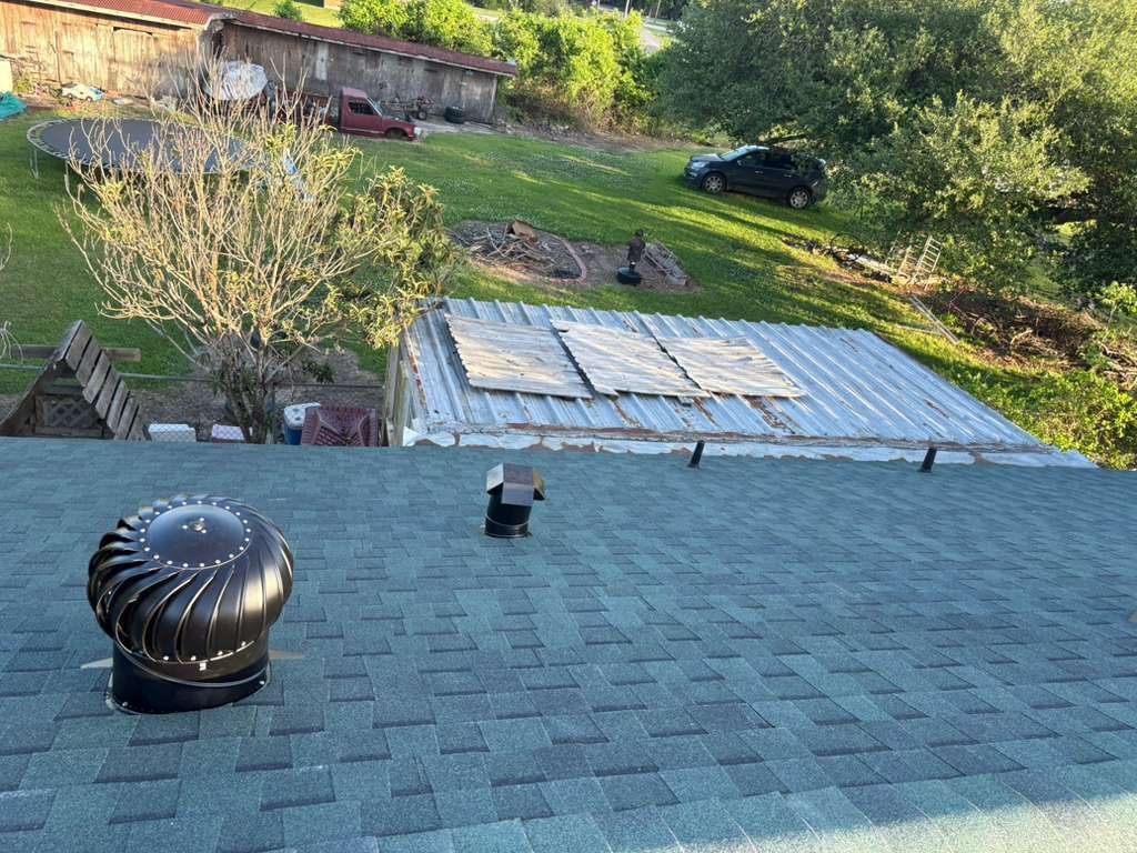Rooftop shingles with a metal vent, overlooking a grassy yard and trees beyond.
